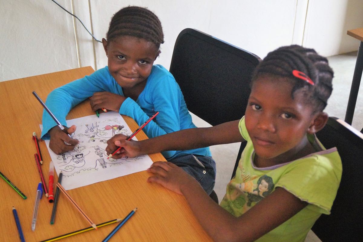 Zwei Mädchen sitzen an einer Schulbank und zeichnen, sehen dabei in die Kamera. © BAS